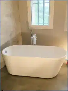 Baignoire îlot blanche moderne avec robinet chromé remplissant la cuve dans une salle de bain aux murs gris béton ciré et fenêtre à barreaux.