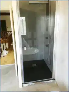 Cabine de douche moderne avec receveur noir, murs gris béton ciré, et lavabo blanc sous plastique. Porte vitrée givrée et fenêtre. Sol clair adjacent.