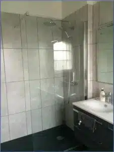 Salle de bain moderne avec douche vitrée, carrelage mural gris clair et sol foncé. Lavabo avec meuble sous vasque sombre.