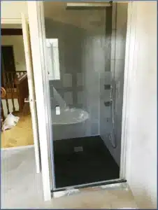 Douche moderne avec receveur noir, vasque ovale, murs gris et fenêtre. Porte vitrée à motifs, sol ciment. Aménagement salle de bain contemporaine.