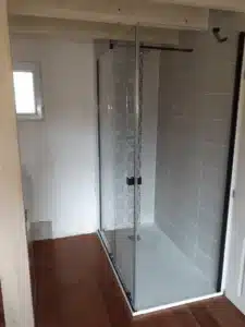 Paroi de douche en verre et profils noirs dans une salle de bain en rénovation. Carrelage gris et motifs, receveur clair, sol bois.
