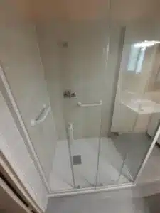 Douche moderne walk-in avec carrelage beige clair, barres d'appui blanches et porte vitrée. Bac à douche extra-plat, drain carré.