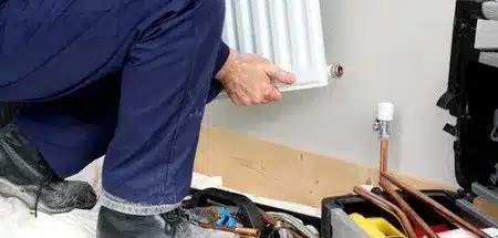 Plombier en vêtements de travail bleus installe un radiateur blanc. Tuyaux de cuivre, vanne et boîte à outils sur sol carrelé.