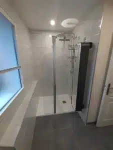 Salle de bain moderne grise avec douche à l'italienne vitrée, grande fenêtre et sèche-serviettes mural noir.