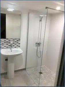 Salle de bain moderne blanche et grise avec lavabo sur pied, miroir éclairé et grande douche à l'italienne carrelée en mosaïque.
