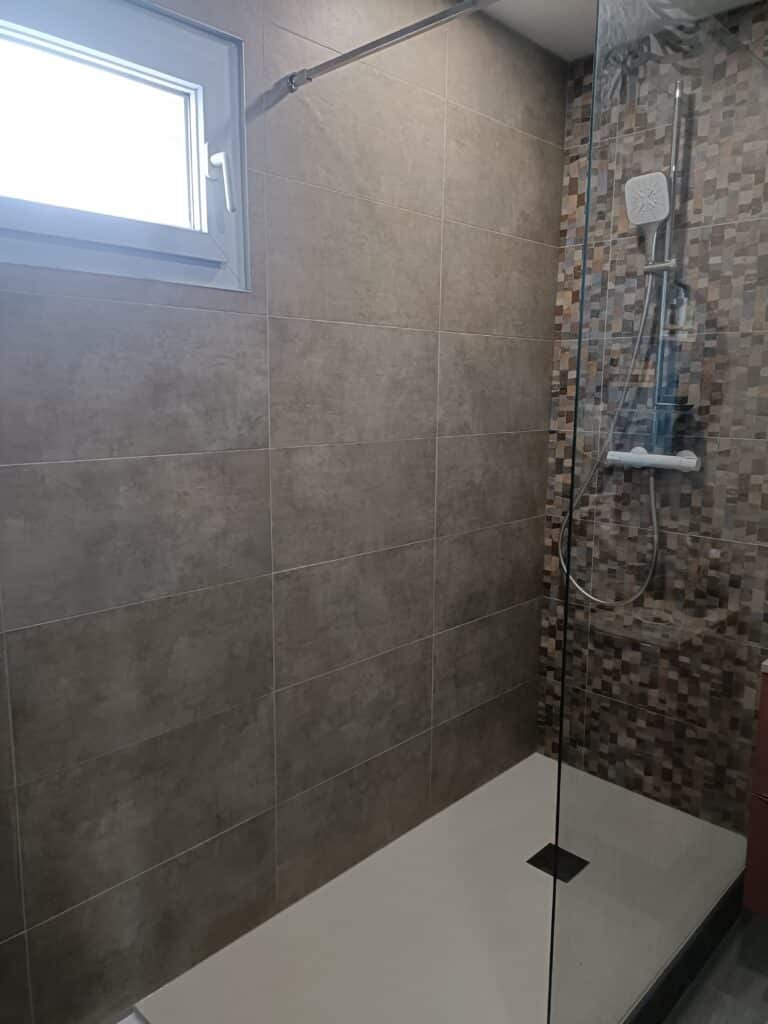 Douche moderne avec grand receveur blanc, carrelage mural gris uni et accent de mosaïque marron près de la pomme de douche carrée. Fenêtre blanche en haut à gauche.
