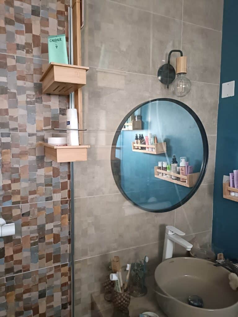 Salle de bain moderne avec murs carrelés, étagères en bois, miroir rond réfléchissant des produits Caudalie, et un lavabo beige.