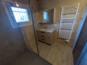 Salle de bain moderne avec meuble-lavabo en bois clair, miroir mural, porte de douche en verre, porte-serviettes chauffant blanc et carreaux beiges.