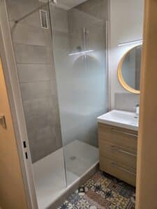 Douche vitrée dépolie et meuble sous-vasque en bois clair dans une salle de bain moderne. Miroir rond éclairé et carrelage de sol à motifs colorés.