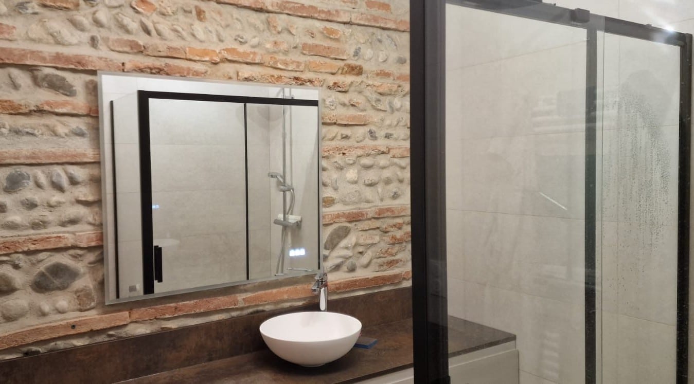 Salle de bain moderne avec mur en pierre naturelle, vasque à poser blanche, miroir et douche à cadre noir.