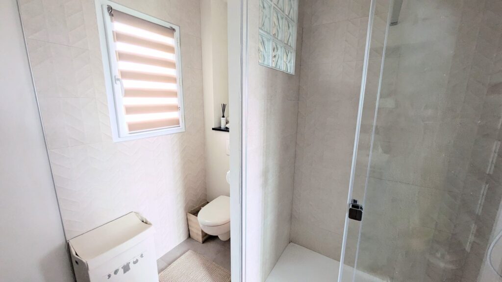Salle de bain moderne avec douche vitrée, WC et carrelage mural beige à motifs chevrons.