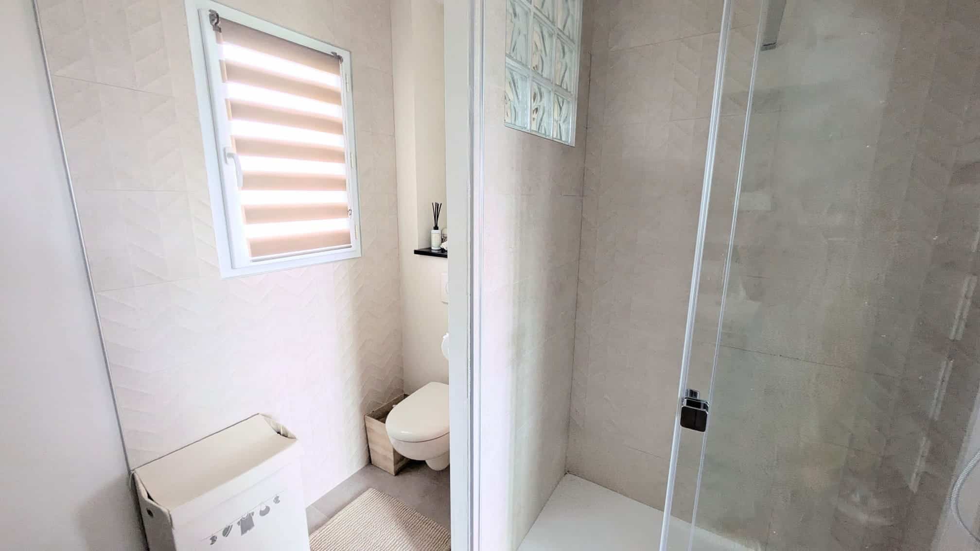 Salle de bain moderne avec douche vitrée, WC et carrelage mural beige à motifs chevrons.