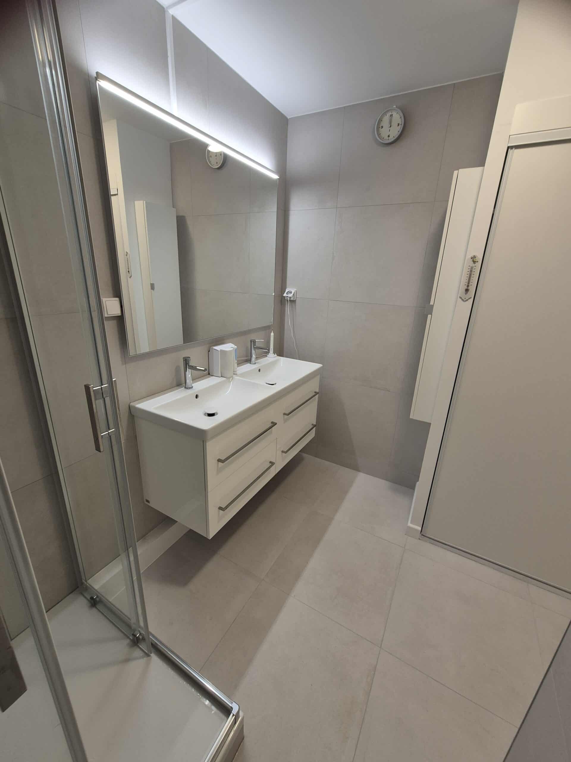 Salle de bain moderne avec double vasque blanche, grand miroir LED et douche vitrée sur carrelage gris.