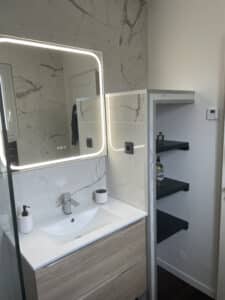 Salle de bain moderne avec miroir LED rétroéclairé, carrelage effet marbre et meuble vasque en bois clair.